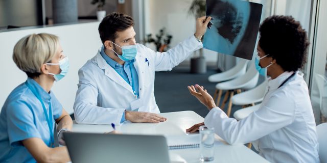 Group of doctors cooperating while analyzing X-ray of a patient at the hospital during coronavirus pandemic. Focus is on male doctor.